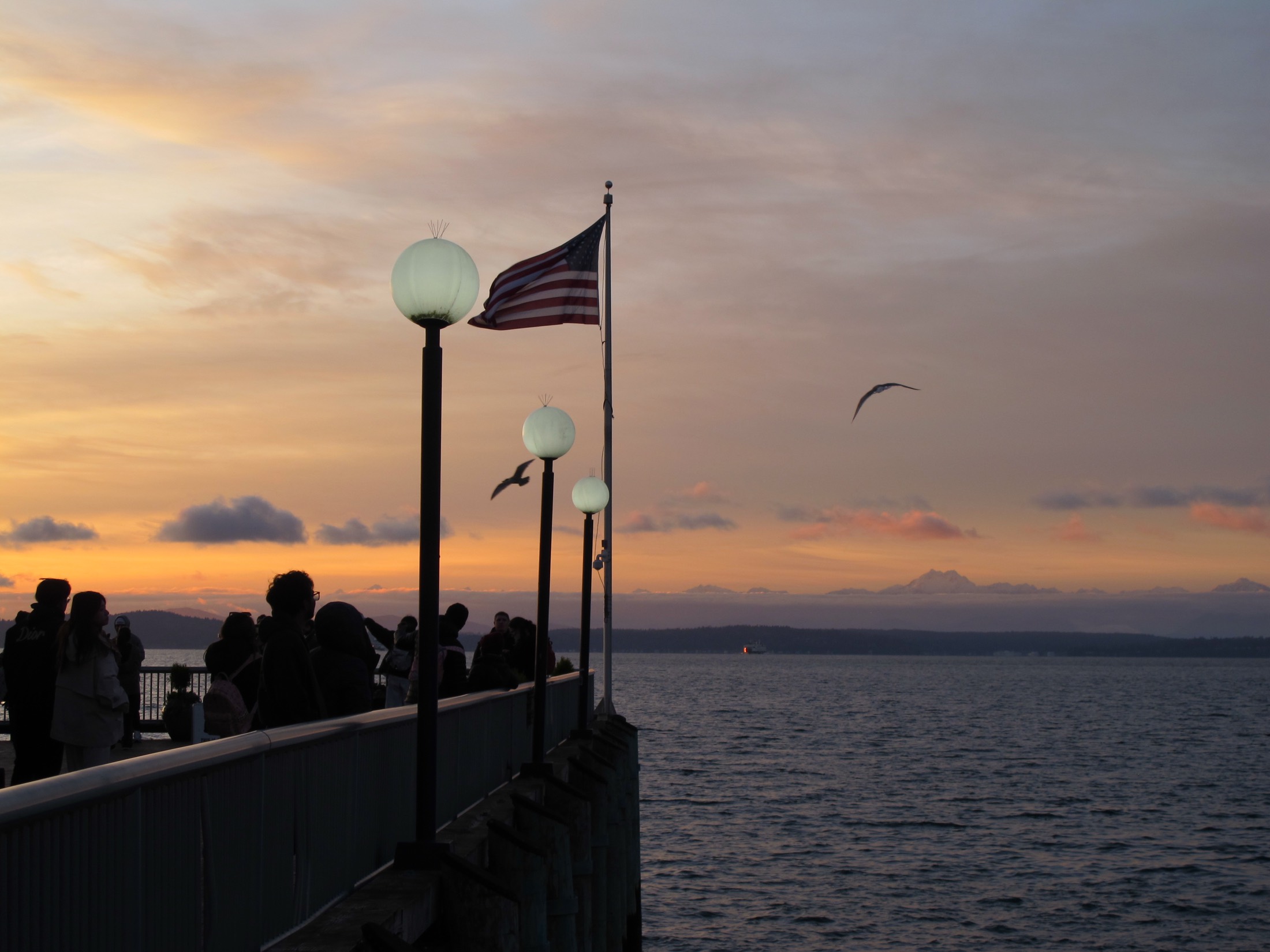 People, gulls, the American flag, and waterfront lamps at sunset over Elliott Bay.